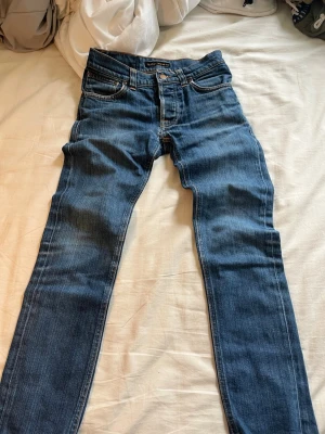 Blå smala jeans från Nudie Jeans - Säljer ett par klassiska blå jeans från Nudie Jeans med smal passform och fem fickor. Jeansen har snygga slitningar och kontrastsömmar samt Nudie-loggan på bakfickan. Perfekta till vardagslooken och tillverkade i mjuk denim.