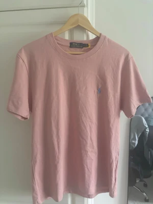 Rosa t-shirt från Polo Ralph Lauren - Säljer en stilren rosa t-shirt från Polo Ralph Lauren med klassisk blå logga broderad på bröstet. T-shirten har rund halsringning och korta ärmar. Tillverkad i mjuk bomull för skön känsla hela dagen.