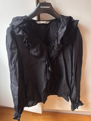 Svart volangblus från ZARA - Svart blus från ZARA med volangdetaljer längs v-ringningen och framtill. Långa ärmar med resår vid ärmslut och lätt skrynkligt tyg. Perfekt för dig som gillar en romantisk och trendig stil.