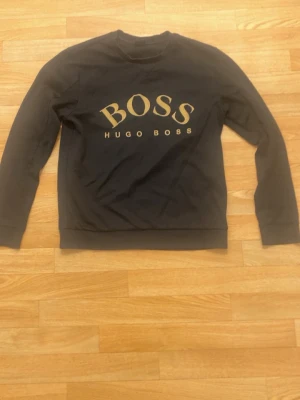 Svart sweatshirt från Hugo Boss - Stilren svart sweatshirt från Hugo Boss med stort guldigt BOSS-tryck på bröstet. Tröjan har rund halsringning, långa ärmar och ribbade muddar vid ärmslut och nederkant. Perfekt för en trendig och avslappnad look.