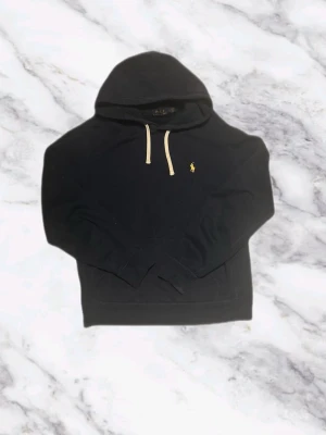 Ralph Lauren Mörkblå Hoodie - Snygg svart hoodie från Polo Ralph Lauren med klassisk gul broderad logga på bröstet och vita dragsnören i huvan. Hoodien har känguruficka framtill och ribbade muddar vid ärmslut och nederkant. Perfekt för en avslappnad och stilren look.