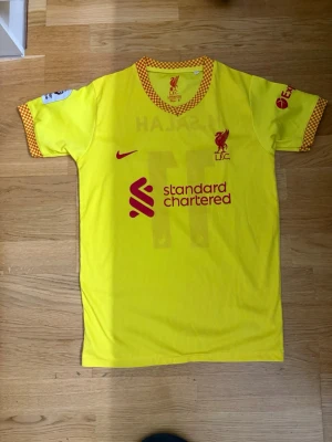 Gul Liverpool fotbollströja Nike - Säljer en gul Liverpool FC fotbollströja från Nike med röda detaljer och rutigt mönster på krage och ärmslut. Tröjan har tryck med M. Salah och nummer 11 på ryggen samt sponsorloggor på bröstet och ärmarna. Perfekt för dig som älskar fotboll och Liverpool!