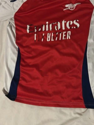 Röd Arsenal matchtröja - Snygg röd och vit Arsenal matchtröja med blå detaljer. Tröjan har tryck med 'Emirates Fly Better' på framsidan och klubbens logga på bröstet. Tillverkad i ett lätt och ventilerande material, perfekt för fotboll eller träning. Men den är inte äckta 