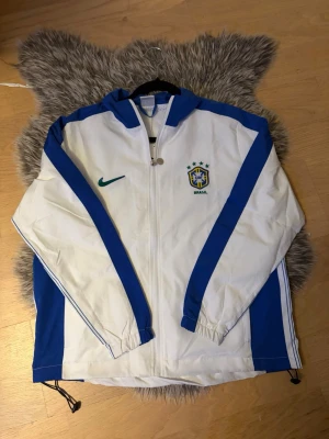 Brasil vindjacka Nike  - Snygg vit windbreaker med blå detaljer och dragkedja. Jackan har Brasilien-logga och Nike-märke på bröstet samt texten 'BRASIL' stort på ryggen. Perfekt för fotbollsfans eller träning. Jackan har resår vid ärmslut och dragsko nertill. Aldrig använd, helt ny. Storlek är S men tycker den sitter som XS. 