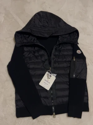 Svart Moncler cardiganjacka med huva - Snygg svart cardiganjacka från Moncler med quiltad front och stickade ärmar. Jackan har en praktisk huva, dragkedja framtill och en ficka med dragkedja på ärmen. Perfekt för lager på lager och streetstyle. Moncler-logga på ärmen och coolt innerfoder med serietidningsmotiv.