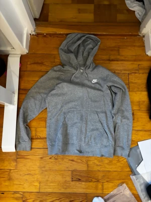 Grå zip-hoodie från Nike - Snygg grå hoodie med dragkedja från Nike. Klassisk modell med huva, två fickor framtill och Nike-logga på bröstet. Perfekt för en avslappnad och sportig stil. Mjuk bomullsblandning och ribbade muddar vid ärmslut och nederkant.