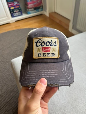 Coors Banquet American Needle  - Retroinspirerad truckerkeps från American Needle med stor broderad Coors Banquet Beer-patch framtill. Mörkgrå front och skärm med beige mesh baktill samt justerbart snapback-spänne.  Kepsen har en tydligt sliten/distressed look på skärmen, vilket är en del av den vintageinspirerade stilen. I övrigt fint begagnat skick.  