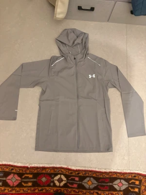 Grå vindjacka från Under Armour - Snygg grå vindjacka från Under Armour med huva och dragkedja framtill. Jackan har vita detaljer på axlarna, två sidofickor och meshfoder för extra komfort. Perfekt för blåsiga dagar och sportiga outfits.