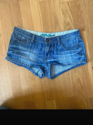 Blå jeansshorts med broderade fickor - Säljer ett par blå denimshorts med låg midja och broderade detaljer på bakfickorna. Tyvärr är dragkedjan lite sönder man kan fortfarande dra upp den men det är lite pilligt. Storlek S/xs Shortsen har klassisk femficksdesign, knappgylf och slitna tvättade partier för en avslappnad look. Perfekta för sommaren!