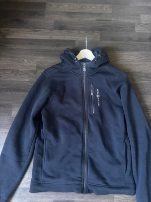 Blå hoodie med dragkedja och huva - Säljer en svart hoodie med hel dragkedja framtill och huva med dragsko. Bröstficka med dragkedja och tryckt text på vänster sida. Två sidofickor och diskret logga på huvan. Perfekt för chill dagar eller när du vill ha en enkel stil.