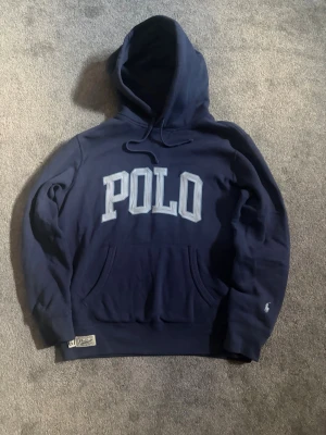 Unik Polo Ralph lauren hoddie  - Snygg mörkblå hoodie från Polo Ralph Lauren med stort POLO-tryck i vitt och ljusblått på bröstet. Klassisk känguruficka framtill, dragsko i huvan och liten broderad logga på ärmen. Perfekt för en avslappnad och trendig stil.