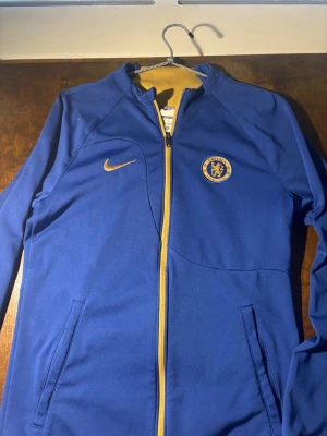 Blå Chelsea Nike träningsjacka - Snygg blå träningsjacka från Nike med Chelsea FC-logga på bröstet och gul Nike-swoosh. Jackan har hel dragkedja i gult, två sidofickor och är i slim fit-modell. Perfekt för fotbollsträning eller som sportig vardagsstil.