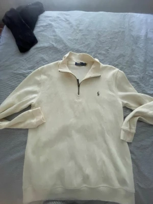 Vit halvzip tröja från Polo Ralph Lauren - Klassisk vit långärmad tröja från Polo Ralph Lauren med halvzip och liten broderad logga på bröstet. Tillverkad i mjukt material och har ribbade muddar vid ärmslut och nederkant. Perfekt för en clean och stilren look.