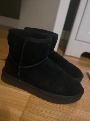 Svarta uggs  - Säljer ett par OANVÄNDA svarta uggs i mjuk mocka med fluffigt foder på insidan. Skorna har en klassisk rund tå och grov sula, perfekta för kyliga dagar. De är ankelhöga och har en enkel, stilren design utan synliga märken.