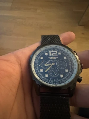 Svart mesh-klocka från Breitling - Stilren klocka från Breitling med svart mesh-armband i metall och stor urtavla. Urtavlan är blå med vita detaljer, stora siffran 12 och tre mindre subdials. Boetten är robust och har en sportig känsla. Perfekt accessoar för dig som gillar klassisk men modern stil.