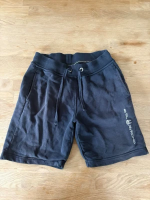 Svarta shorts med snörning Sail Racing - Svarta shorts från Sail Racing med snörning i midjan och tryckt logga på benet. Modellen har en relaxed fit och är tillverkad i mjuk bomull, perfekta för sommardagar eller chill hemma.