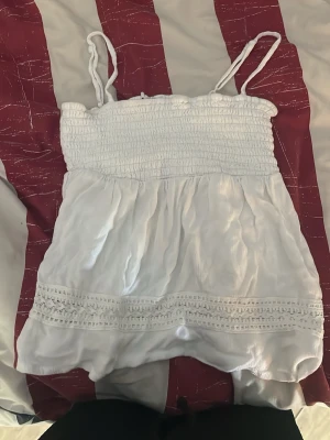 Vit smockad sommartopp med spets - Söt vit topp med smock upptill och tunna axelband. Toppen har ett luftigt fall och dekorativ spetsdetalj nedtill. Perfekt för varma dagar och enkel att matcha med jeans eller kjol.
