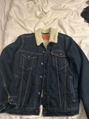 Mörkblå Levis jeansjacka  - Helt ny oanvänd perfekt skick varm o skön. Snygg mörkblå jeansjacka med vit teddykrage och teddyfoder. Klassisk design med två bröstfickor med lock, knappar framtill och kontrastsömmar. Perfekt för dig som vill ha en cool och tidlös look.