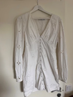 Vit broderad klänning med knappar - Söt vit klänning med broderade blommor och dekorativa knappar framtill. Klänningen har långa ärmar, v-ringning och är gjord i ett lätt tyg som känns luftigt. Perfekt för vår och sommar.