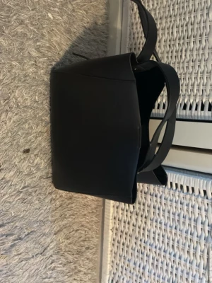 Svart stilren handväska i skinnimitation - Snygg svart handväska med minimalistisk design och bred axelrem. Väskan är tillverkad i skinnimitation och har ett rymligt huvudfack samt innerficka med dragkedja. Perfekt för dig som gillar enkel och modern stil.