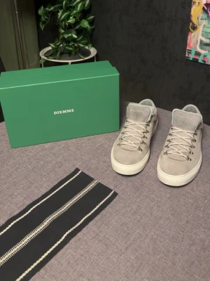 Gråa Diemme sneakers i mocka - Snygga gråa sneakers från Diemme i mjuk mocka med vita sulor och metallöglor för snörningen. Klassisk låg modell med stilren design och diskret logga på plösen. Perfekta för dig som gillar minimalistisk och clean stil.
