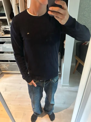Mörkblå tröja med rund hals - En stilren mörkblå tröja med rund hals och diskret logga på bröstet. Tröjan har långa ärmar och ribbade muddar vid ärmslut och nederkant. Perfekt för en clean och enkel look.