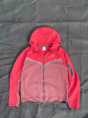 Vin röd nike tech fleece - Säljer en röd hoodie från Nike med hel dragkedja framtill och huva. Hoodien har två fickor, svarta detaljer och en liten ficka med dragkedja på ena ärmen. Tillverkad i mjukt bomullsmaterial och har Nike-logga på bröstet.