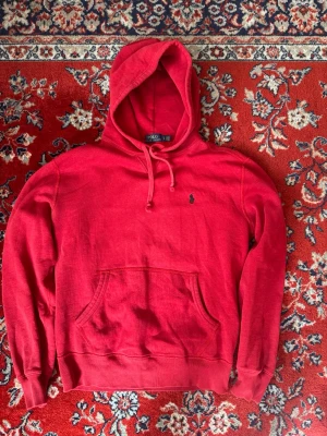 Röd hoodie från Polo Ralph Lauren - Säljer en snygg röd hoodie från Polo Ralph Lauren med klassisk huva och dragsko. Hoodien har en känguruficka framtill och den ikoniska lilla Polo-loggan broderad på bröstet. Tillverkad i mjukt bomullsmaterial och har ribbade muddar vid ärmar och nederkant.