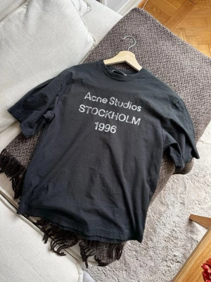 Svart Acne Studios t-shirt - Svart t-shirt från Acne Studios med vit text 'Acne Studios STOCKHOLM 1996' på bröstet. Klassisk rund halsringning och korta ärmar. Tillverkad i bomull och har en loose passform som ger en avslappnad vibe.