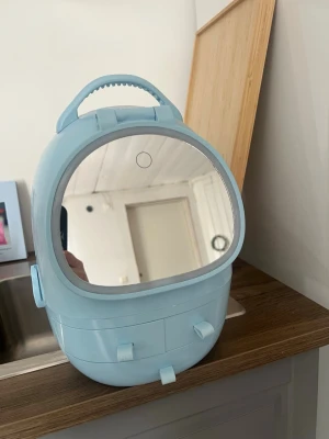 Blå sminkspegel med förvaring - Snygg sminkspegel i ljusblå plast med rundad form och praktiskt handtag. Spegeln har inbyggd LED-belysning och flera förvaringsfack, inklusive utdragbara lådor. Perfekt för att organisera smink och accessoarer hemma eller på resan.