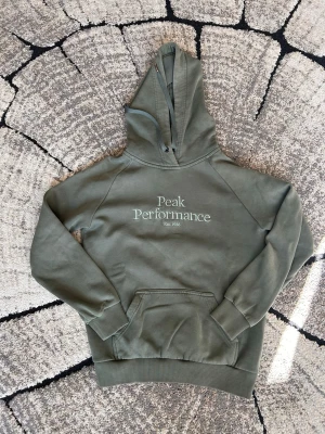 Olivgrön hoodie Peak Performance - Snygg olivgrön hoodie från Peak Performance med broderad logga och text 'Est. 1986' på bröstet. Klassisk känguruficka framtill, ribbade muddar och justerbar huva med dragsko. Tillverkad i mjuk bomullsblandning som känns skön mot huden.