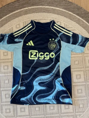 Ajax blå matchtröja Adidas - Snygg blå Ajax matchtröja från Adidas med ljusblå och gula detaljer. Tröjan har korta ärmar, rund hals och ett vågigt mönster framtill. Ajax klubbmärke och Ziggo-logga på bröstet samt tre gula ränder på axlarna. Perfekt för fotbollsträning eller match. Passar dig som är 160-170 cm lång. 