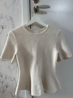 Vit/beige kortärmad stickad tröja med peplum-effekt från H&M – Ny och Storlek 38/M/10 - Snygg ribbad topp i ljus stickad kvalitet med korta ärmar och rund hals. Modellen har en lätt utsvängd passform som ger en feminin siluett. Perfekt att styla med jeans eller kjol för en trendig look.