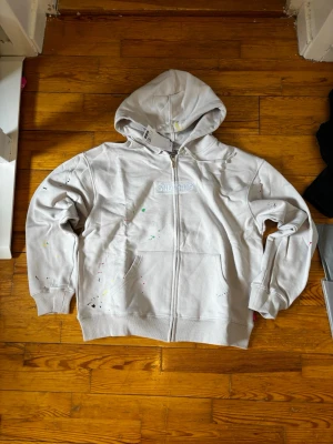 Vit Supreme x Margiela zip hoodie - Säljer en vit zip hoodie från Supreme i samarbete med Maison Margiela. Hoodien har huva, dragkedja framtill, fickor och diskreta färgstänk över hela plagget. Supreme-logga broderad på bröstet. Perfekt för dig som gillar streetwear och unika detaljer.