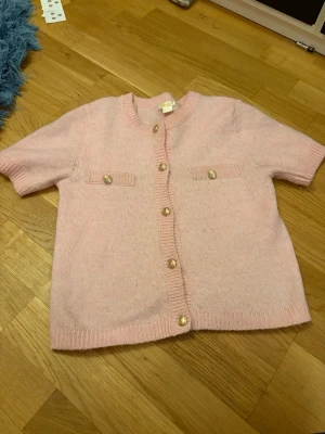 Ljusrosa stickad kofta Gina Tricot - Supersöt ljusrosa stickad kofta från Gina Tricot med korta ärmar och rund hals. Koftan har dekorativa guldknappar framtill och två små fickor på bröstet. Perfekt att slänga över en topp för en gullig look.