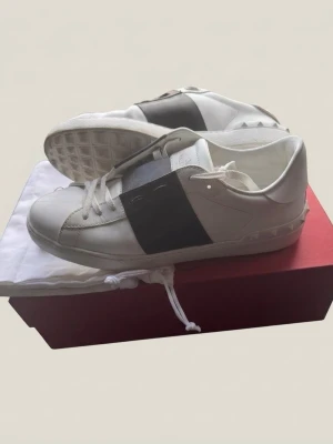 Valentino Open St. 43 - Stilrena vita sneakers med bred svart rem över mitten. Skorna har klassisk låg profil, vita snören och sula med mönster. Tillverkade i slätt läder och har en modern, minimalistisk look.