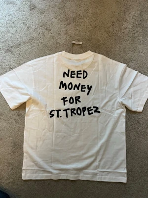 Vit t-shirt med tryck  - Säljer en vit t-shirt från Only & Sons med svart texttryck 'NEED MONEY FOR ST. TROPEZ' både fram och bak. T-shirten har rund hals och korta ärmar. Perfekt för dig som gillar statement-plagg och enkel stil.