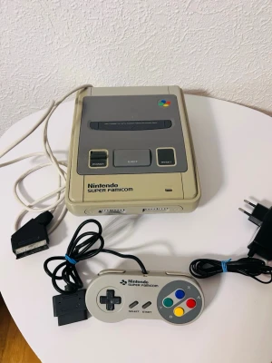 Nintendo Super Famicom konsol - Klassisk Nintendo Super Famicom spelkonsol i grått med tillhörande handkontroll och kablar. Konsolen har rundade former och färgglada knappar på kontrollen. Perfekt för retrospel och nostalgi med vänner.