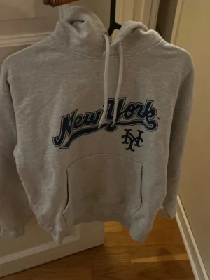 Grå New York Mets hoodie - Säljer en grå hoodie med New York Mets-tryck i blått framtill. Klassisk känguruficka, huva med vita snören och ribbade muddar. Perfekt för dig som gillar sportig streetstyle och vill ha en bekväm och snygg tröja till vardags.    Mått: 48 cm bröst / 55 cm längd / 48 cm ärm.