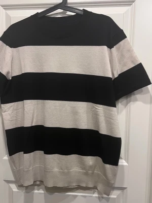 Randig svart och beige t-shirt blus - Säljer en stilren t-shirt med breda svarta och beige ränder. T-shirten har rund halsringning och korta ärmar. Materialet känns mjukt och skönt, troligtvis bomull eller en bomullsblandning. Perfekt för en avslappnad look.