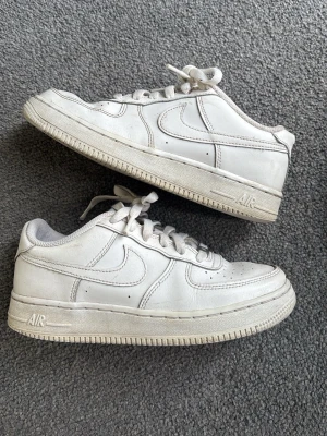 Vita klassiska sneakers i läder - Säljer ett par vita Nike Air Force 1 sneakers i lågt modell. Skorna är i läder med perforerade detaljer på tån och har den ikoniska swoosh-loggan på sidan. Tjock sula med AIR-detalj och klassisk snörning. Perfekta för en clean och stilren look.