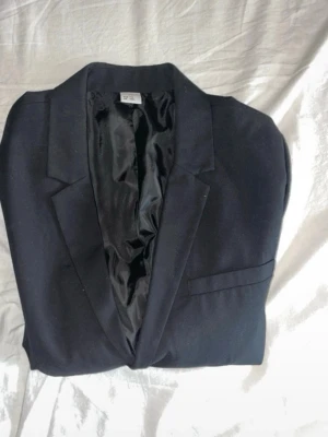 Svart kavaj från Lindex Kids - Stilren svart kavaj från Lindex Kids i storlek 170. Klassisk modell med spetsiga slag, helsvart foder och enkel bröstficka. Perfekt för dig som vill ha en clean och tidlös look.