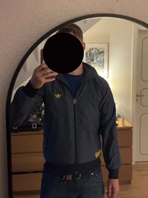 Adidas vindjacka  - Snygg blå vindjacka från Adidas Chile 62-kollektionen med gul logga på bröstet och detaljer vid fickan. Jackan har hel dragkedja, två sidofickor och svarta muddar vid ärmslut och midja. Materialet är lätt och passar perfekt till streetwear.