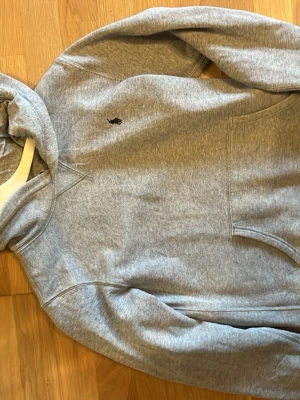 Grå hoodie från Polo Ralph Lauren - Snygg grå hoodie från Polo Ralph Lauren med klassisk huva och känguruficka. Tröjan har det ikoniska lilla Polo-märket broderat på bröstet och är gjord i mjukt bomullsmaterial. Perfekt för en avslappnad och stilren look.
