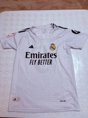 Real Madrid (Valverde 15) - Snygg vit Real Madrid matchtröja från Adidas med Valverde och nummer 15 på ryggen. Tröjan har svarta detaljer, klubbmärke på bröstet och sponsortryck. Tillverkad i lätt och ventilerande material, perfekt för fotbollsträning eller match.