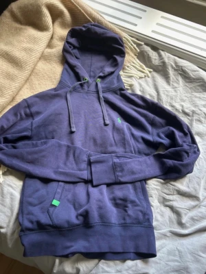Ralph Lauren - Snygg lila hoodie med huva och dragsnören, samt gröna detaljer på snörhålen och en liten grön logga på bröstet. Känguruficka framtill och ribbade muddar vid ärmar och nederkant. Perfekt för en avslappnad och trendig look.