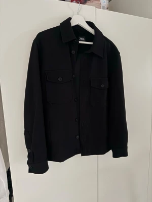 Svart overshirt från ZARA -  Svart overshirt från ZARA med klassisk krage, raka ärmar och två stora bröstfickor med lock och knapp. Skicket är som nytt 10/10. Overshirten har knäppning framtill och vid ärmslut, perfekt för lager på lager. Materialet är mjukt och lite tjockare, vilket ger en snygg och avslappnad look.