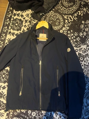 Mörkblå Moncler jacka med dragkedjor - Snygg mörkblå jacka från Moncler med ståkrage och två vertikala dragkedjefickor framtill. Jackan har en diskret Moncler-logga på vänster ärm och är tillverkad i ett slätt, lätt material. Perfekt för dig som vill ha en stilren och exklusiv look.