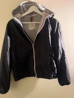 Svart vindjacka med huva från Moncler - Snygg svart vindjacka från Moncler med grått innerfoder och huva. Jackan har dragkedja framtill, två sidofickor med knappar och Moncler-logga på ärmen. Perfekt för blåsiga dagar och enkel att matcha till olika outfits.