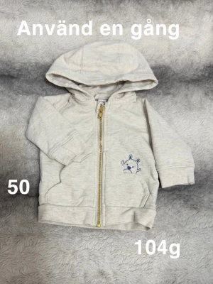 Creamfärgad hoodie med dragkedja, stl 50 - Supersöt creamfärgad hoodie med huva och dragkedja framtill. Har en liten blå björn-illustration på ena sidan och två fickor. Perfekt för mysiga dagar och enkel att ta på tack vare dragkedjan. Mjuk och bekväm, passar de allra minsta.🌸 Använd en gång. Storlek 50